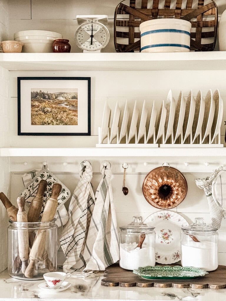 A cozy, vintage-style kitchen with open shelves displaying plates, jars, rolling pins, baskets, a scale, a framed landscape photo, dish towels, and a copper mold hanging on a white wall.