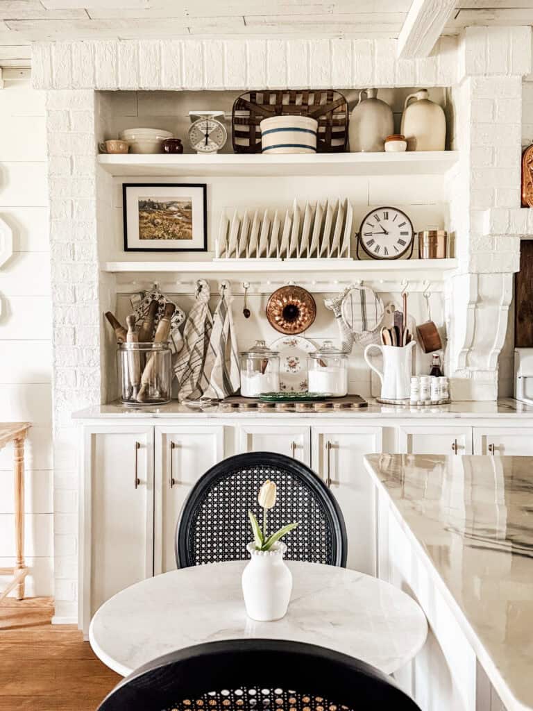 A cozy, white farmhouse kitchen with open shelves displaying dishes, jars, and decor. A small round table with a marble top and a vase with a single tulip sits in front of black chairs. Dishes and utensils are neatly organized.