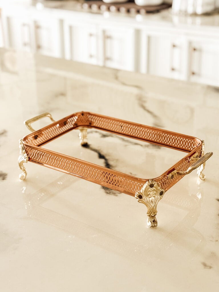 A decorative rectangular tray with ornate gold handles and legs, featuring a copper-colored mesh border, sits on a shiny marble countertop in a bright kitchen.
