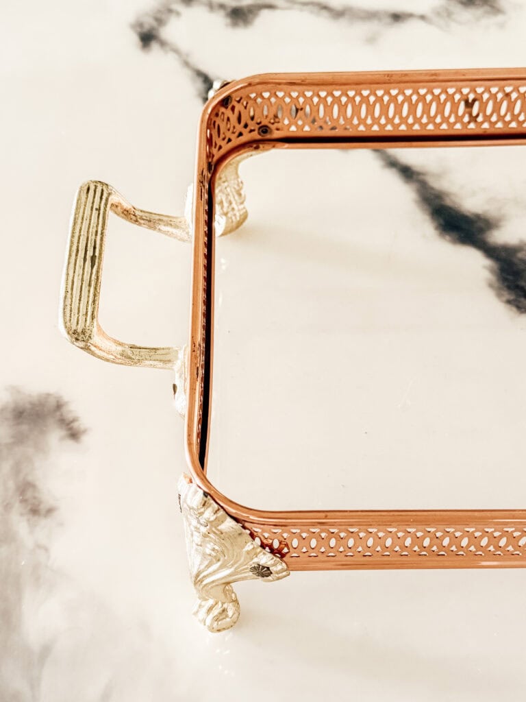 A close-up of a decorative rectangular tray with ornate gold handles and legs, and a perforated copper rim, placed on a marble surface with dark, shadowy patterns.