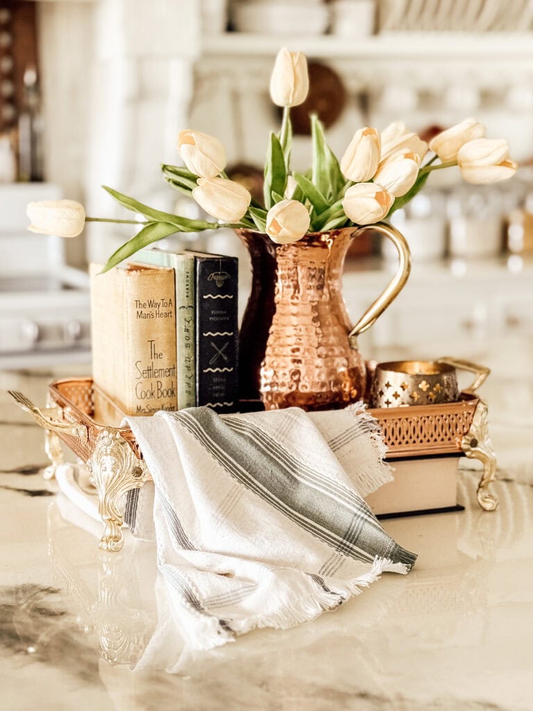 A decorative tray holds a copper pitcher with pale tulips, two vintage cookbooks, a candle, and a folded white cloth with green stripes, all displayed on a marble countertop in a cozy kitchen setting.