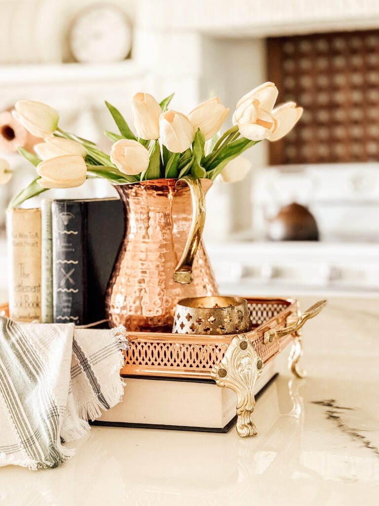 A copper pitcher holding cream-colored tulips sits on a decorative tray with books, a candle holder, and a striped cloth, placed on a bright kitchen countertop.