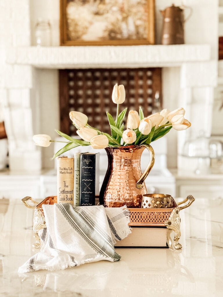 A decorative tray holds a hammered copper pitcher with white tulips, two books, and a folded striped cloth on a shiny surface. A fireplace and a framed painting are in the blurred background.