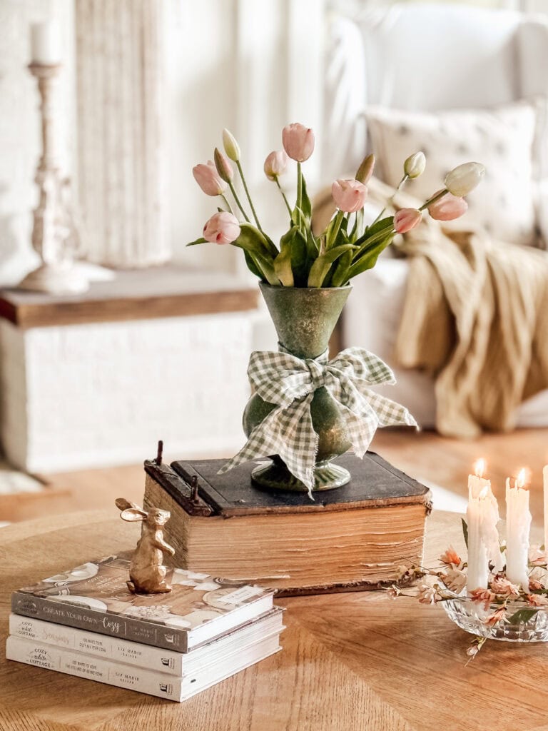 A vase with pink tulips and a gingham ribbon sits on a stack of vintage books on a round wooden table. Nearby are three lit candles, more books, and a small decorative rabbit, with cozy decor in the background.