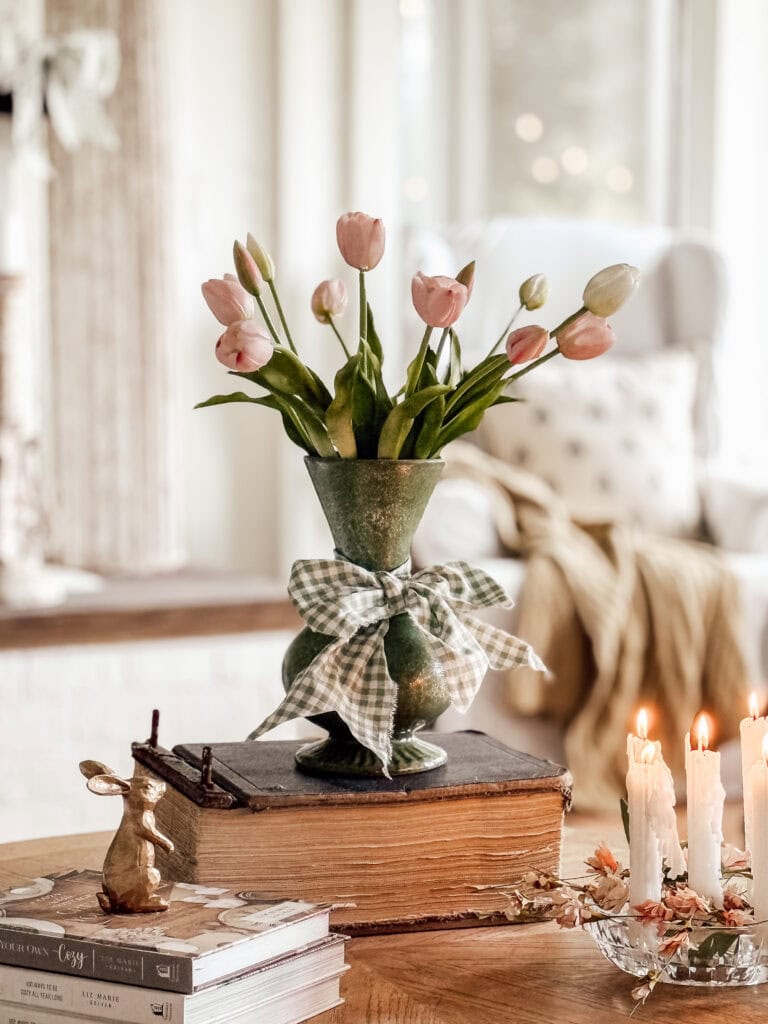 A green vase with pink tulips and a plaid bow sits on old books, next to three lit candles and a small rabbit figurine, all on a wooden table in a cozy, softly lit living room.