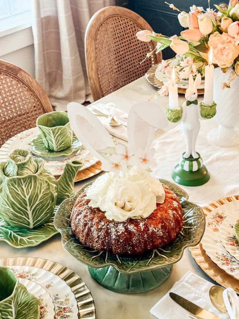 A Bundt cake topped with white flowers and bunny ears sits on a green leaf-shaped cake stand, surrounded by elegant tableware, floral plates, cabbage-shaped dishes, and a spring-themed centerpiece.