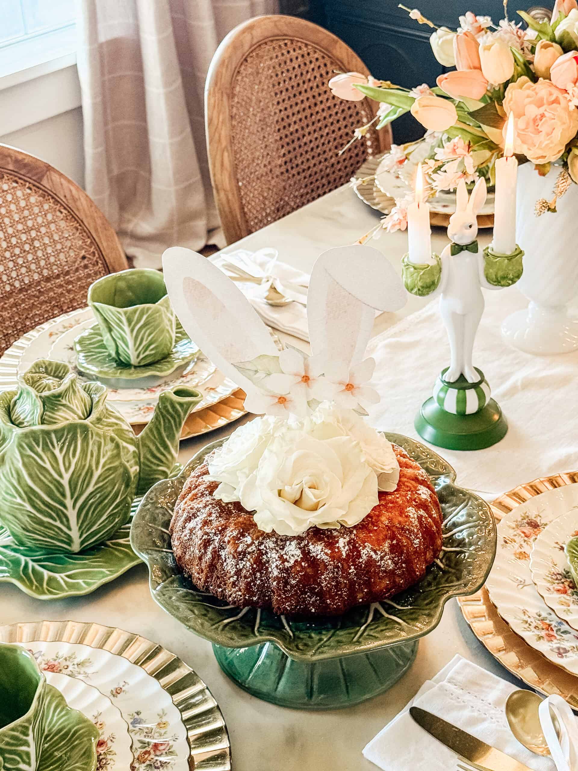 A Bundt cake topped with white flowers and bunny ears sits on a green leaf-shaped cake stand, surrounded by elegant tableware, floral plates, cabbage-shaped dishes, and a spring-themed centerpiece.