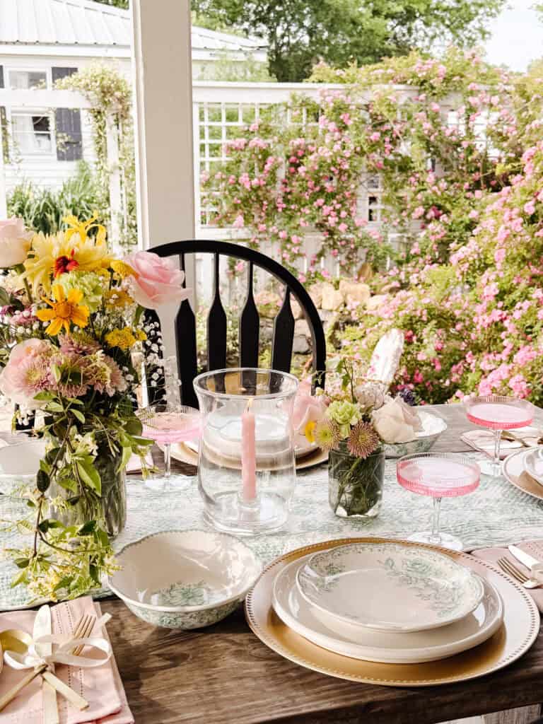 A beautifully set outdoor table features floral plates, gold cutlery, pink glasses, and vases of fresh flowers. The background shows lush pink roses and greenery, creating a charming garden atmosphere.