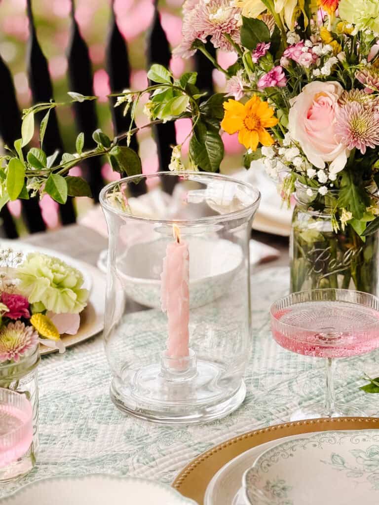 A pastel pink candle burns inside a glass hurricane lantern on a decorated table set with floral plates, pink drinks, and colorful flower arrangements in jars. Pink flowers and greenery appear in the background.