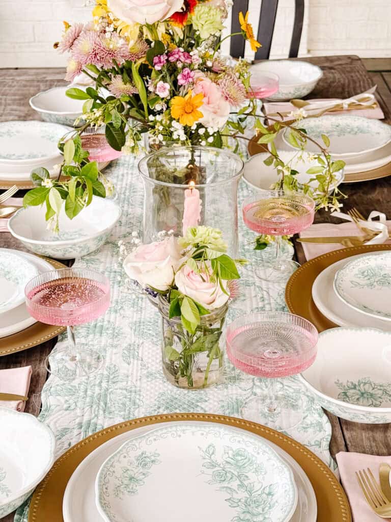 A beautifully set dining table features floral plates, gold chargers, pink napkins, pink drinks in coupe glasses, and vases with roses and assorted flowers, creating an elegant and festive atmosphere.