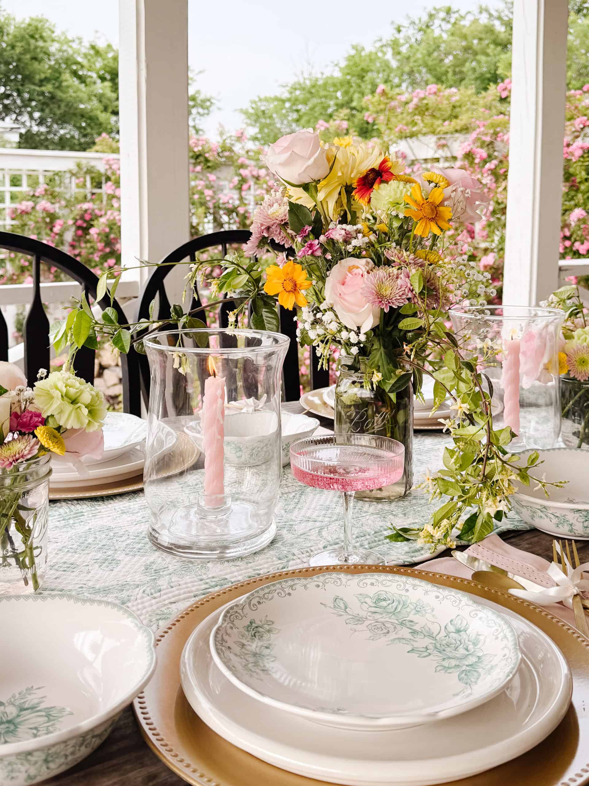A spring-themed outdoor table is set with floral plates, gold chargers, glassware, pastel pink candles, and a centerpiece of pink and yellow flowers, with blooming bushes in the background.