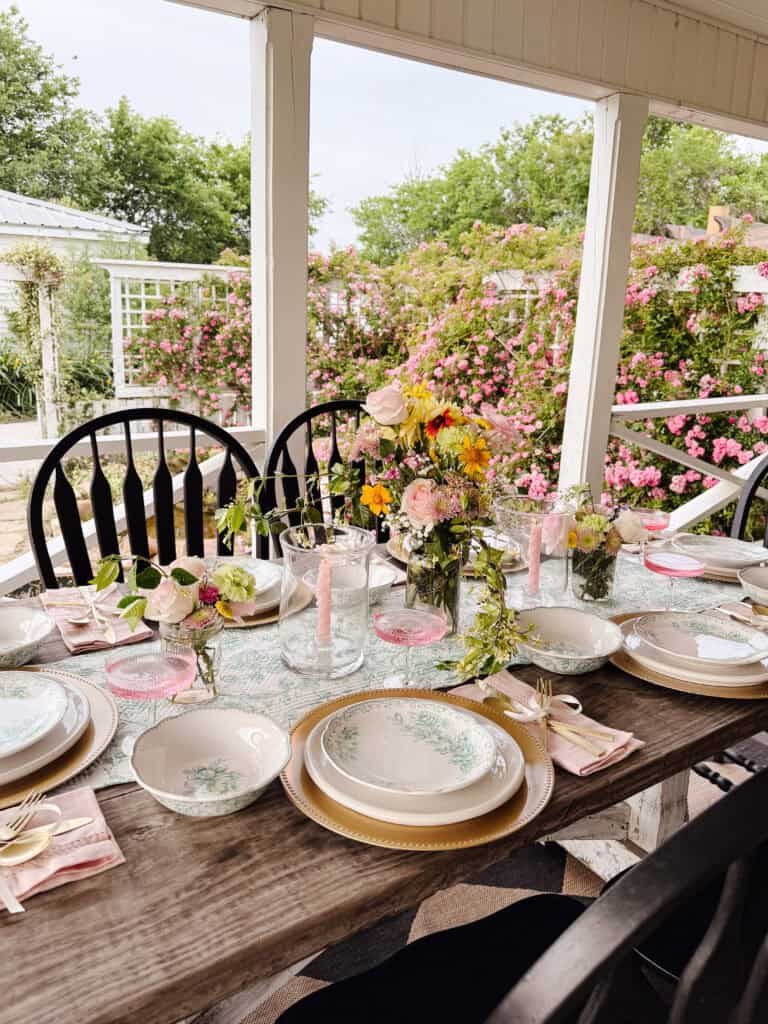 A beautifully set outdoor dining table features floral plates, gold-rimmed glasses, pink candles, and small flower arrangements. The table sits on a porch with blooming pink flowers in the background.