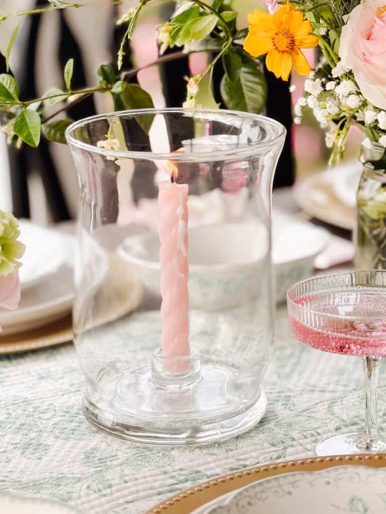 A pink twisted candle burns inside a clear glass hurricane holder on a floral-patterned tablecloth, surrounded by elegant tableware and colorful flower arrangements.