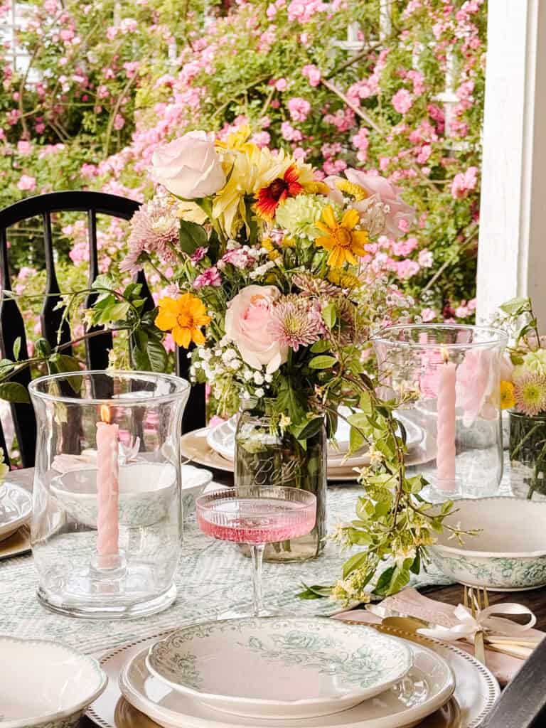 A beautifully set table featuring floral-patterned plates, a pink drink in a glass, pink twisted candles in glass holders, and a vase of colorful flowers, with blooming pink roses in the background.