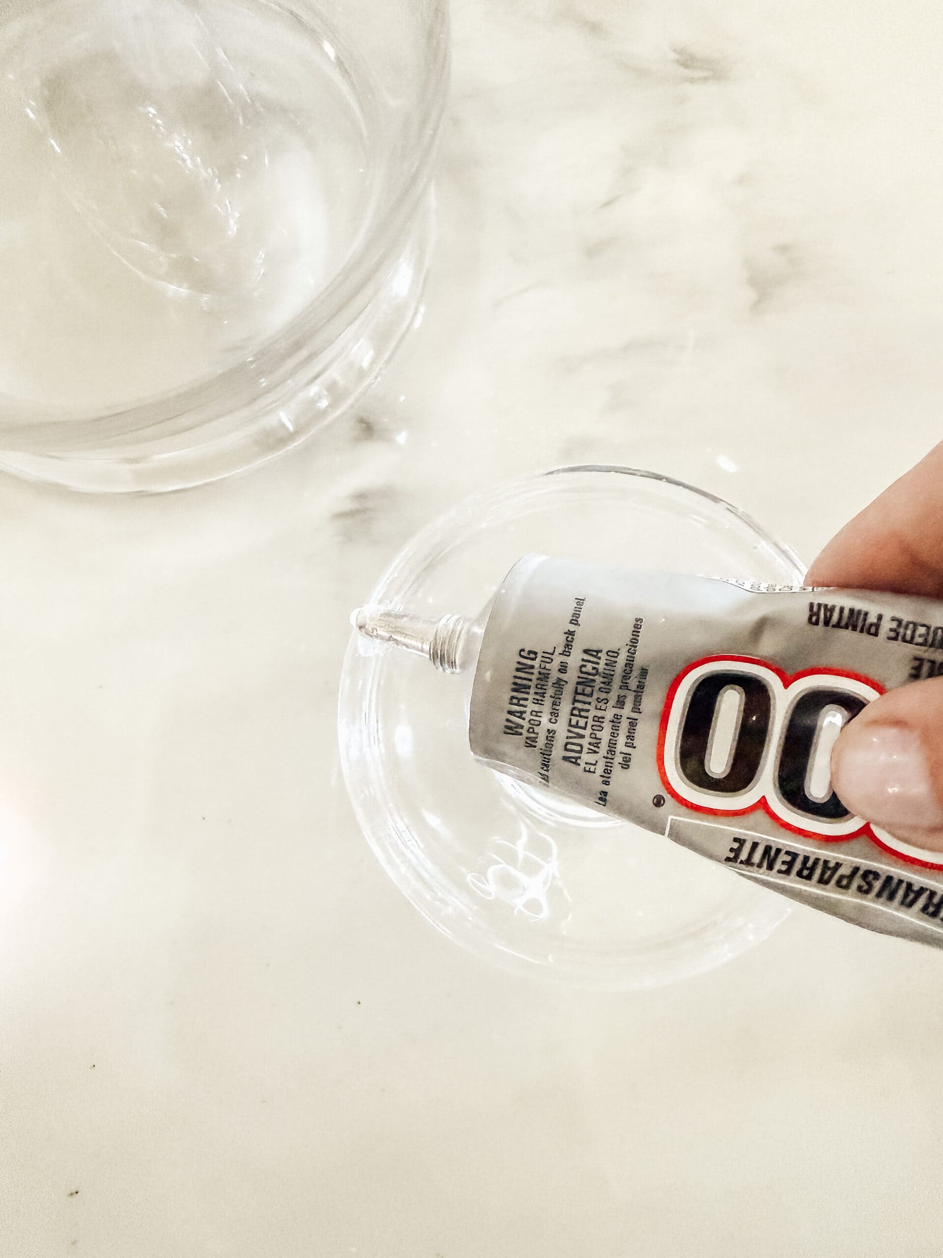 A hand squeezing clear adhesive glue from a tube labeled E6000 into a small glass bowl on a white marble surface. Another empty glass bowl is nearby.