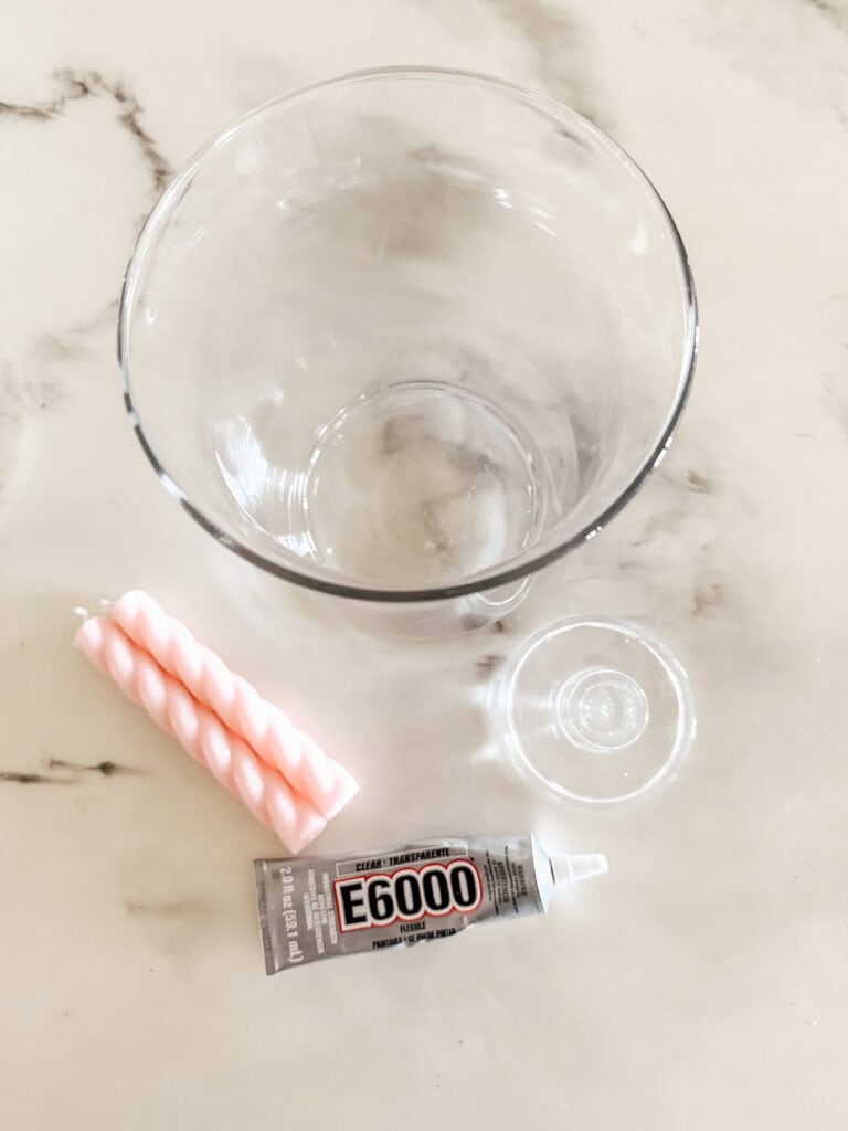 A clear glass cup, a small glass base, a pink twisted candle, and a tube of E6000 glue are arranged on a white marble surface.