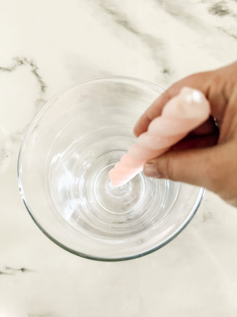 A hand holds a pink, twisted candle and dips its tip into a clear glass bowl filled with water, on a white marble surface.