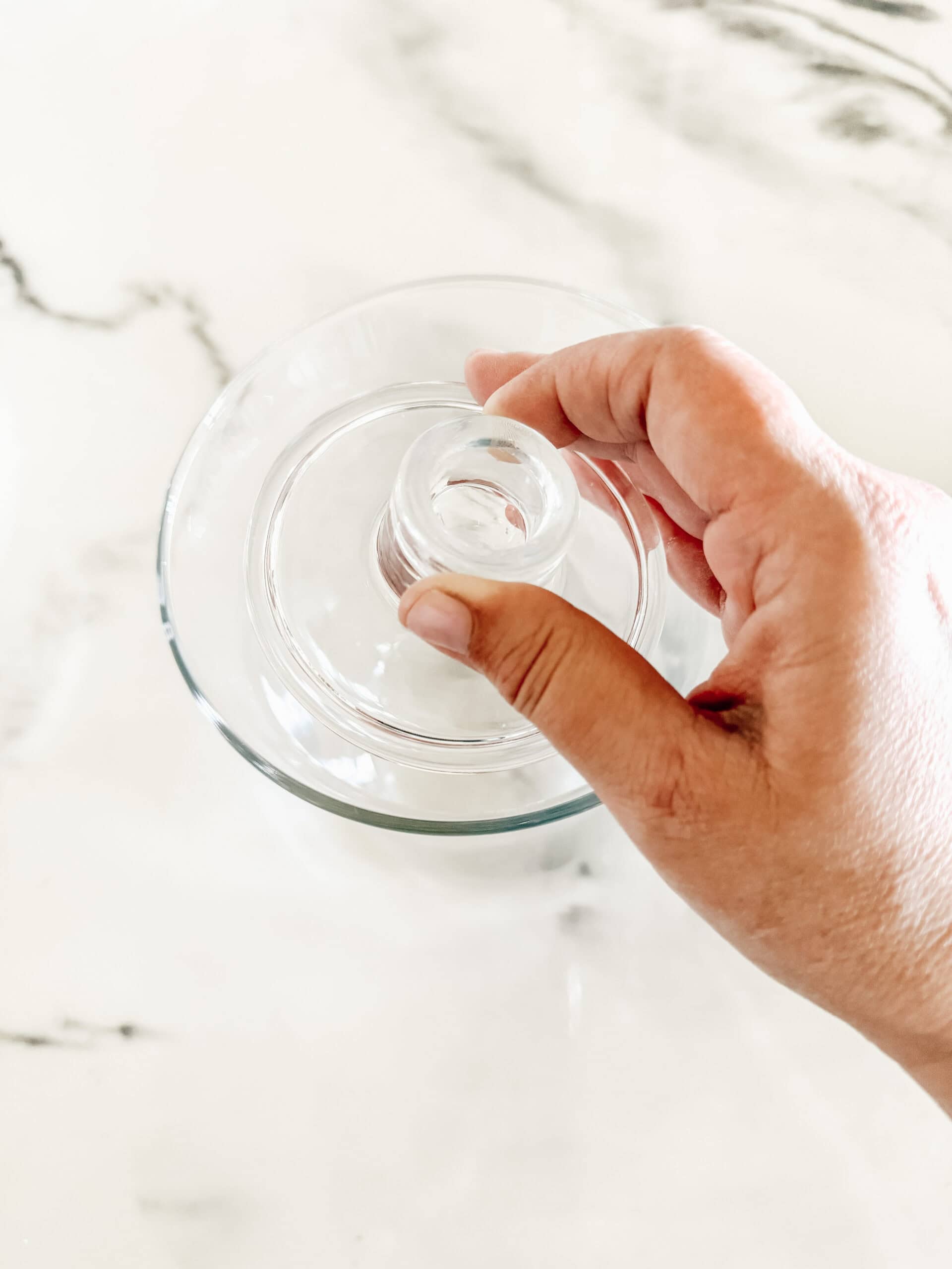 A hand is holding a glass lid above a clear glass bowl on a white marble surface.