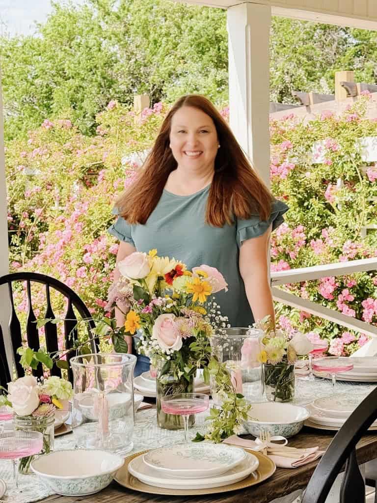 A woman with long brown hair stands smiling behind a beautifully set outdoor table with floral arrangements, glassware, and plates. Pink flowers and greenery fill the background on a sunny day.