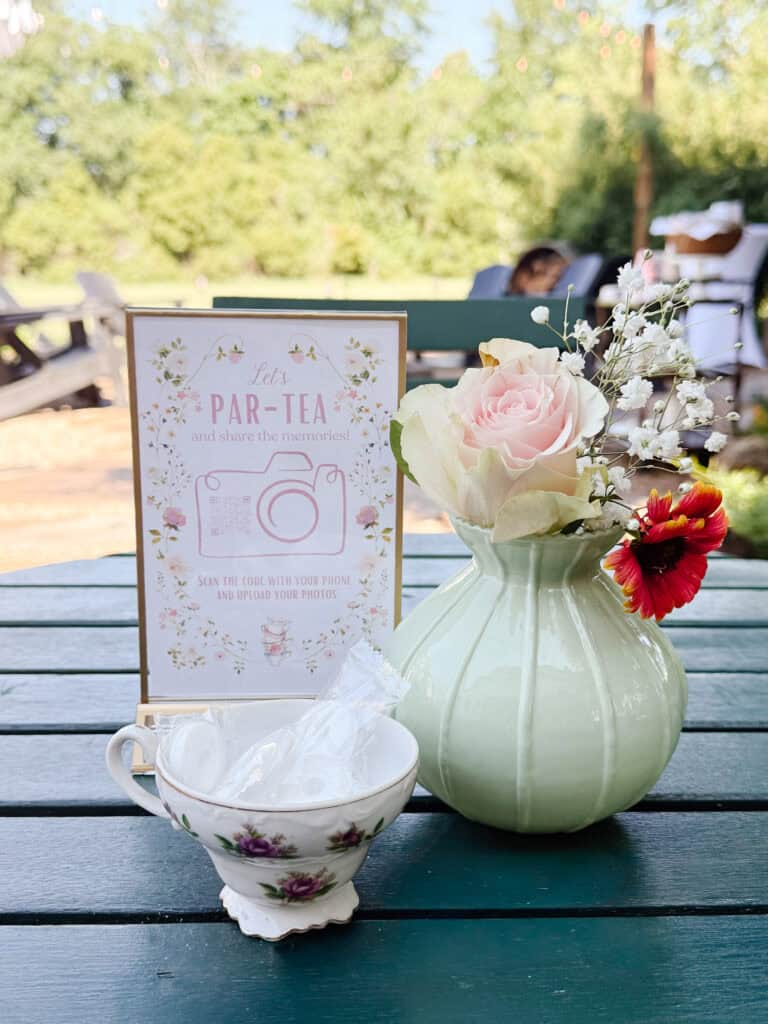 A pastel green vase with roses and small flowers sits on a teal table next to a teacup with wrapped candies and a floral sign that reads Lets Par-Tea in an outdoor garden setting.