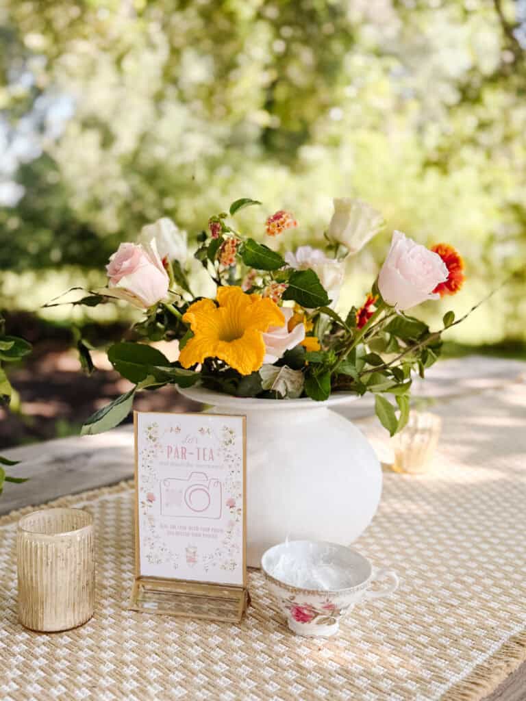 A white vase with colorful flowers sits on a woven tablecloth outdoors. Next to it are a vintage teacup, a small card with floral designs, and a candle holder, all under dappled sunlight with greenery in the background.