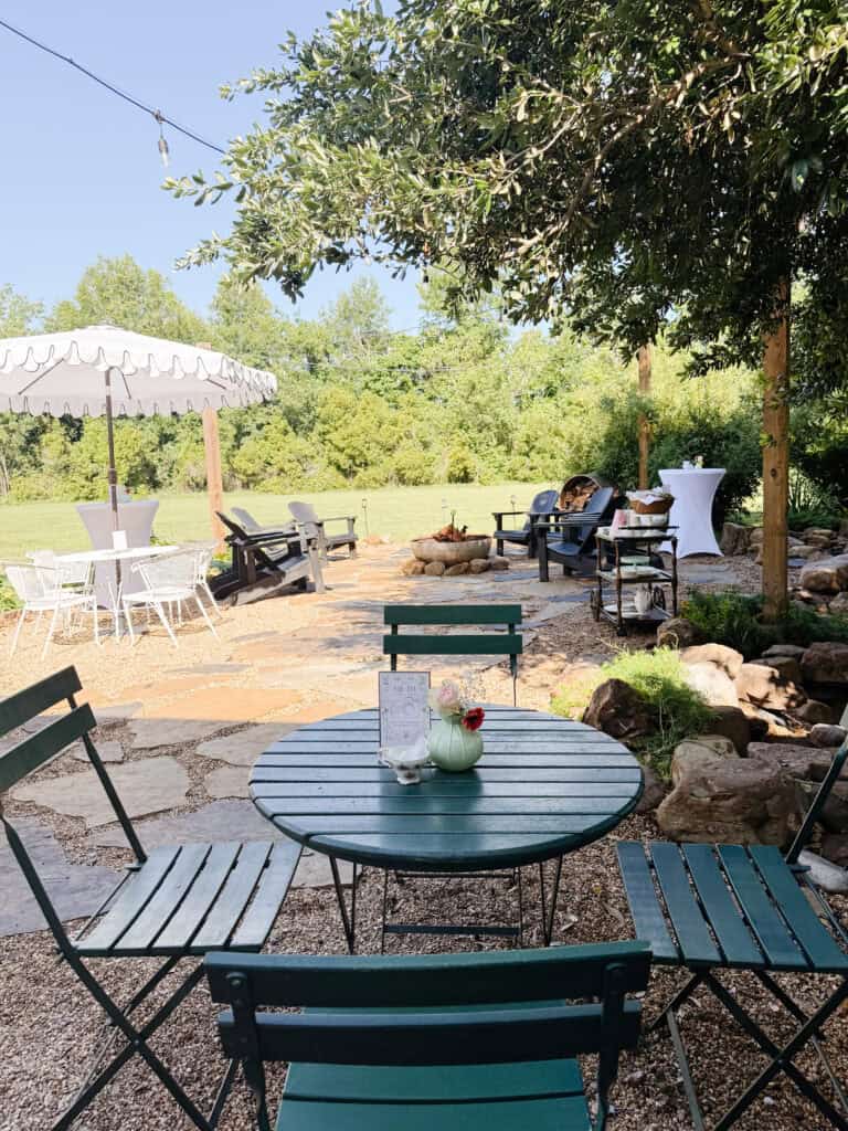 Outdoor cafe scene with green metal table and chairs, potted plant centerpiece, stone patio, trees, string lights, white umbrella, and relaxed seating in a lush, sunny garden setting.
