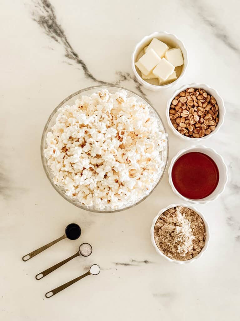 A bowl of popcorn surrounded by small bowls containing butter, peanuts, red sauce, brown sugar, and three measuring spoons with black, white, and gray powders, all on a light marble surface.
