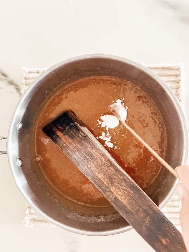 A hand sprinkles white powder from a small stick into a pot of brown liquid on a woven mat, while a large wooden spatula rests inside the pot.