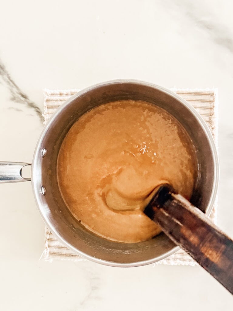 A saucepan containing a light brown, thick mixture is being stirred with a wooden spoon, placed on a woven mat atop a light marble surface.