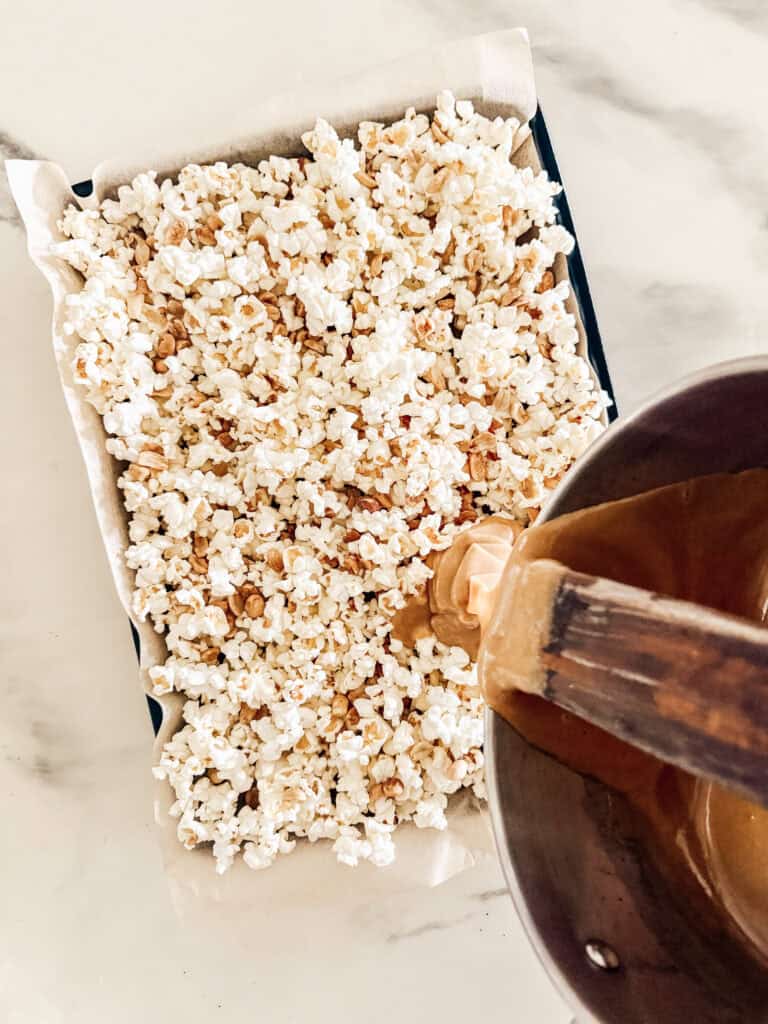 A baking tray lined with parchment paper and filled with popcorn sits on a counter while caramel is being poured over it from a saucepan.