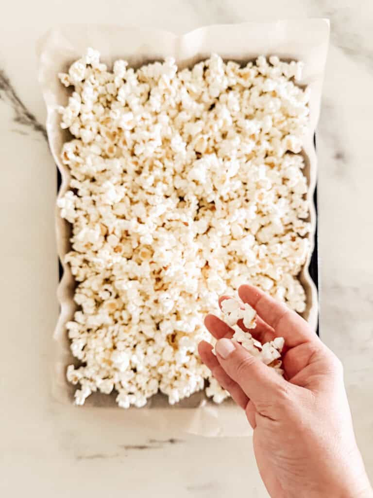 A hand picking up popcorn from a baking tray lined with parchment paper, filled with freshly popped popcorn on a light-colored surface.