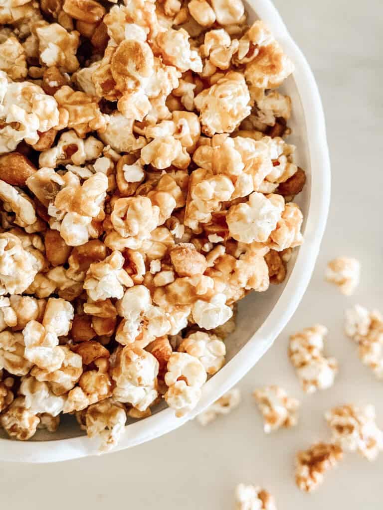 A close-up of a white bowl filled with caramel-coated popcorn, with a few pieces scattered on a light-colored surface beside the bowl.