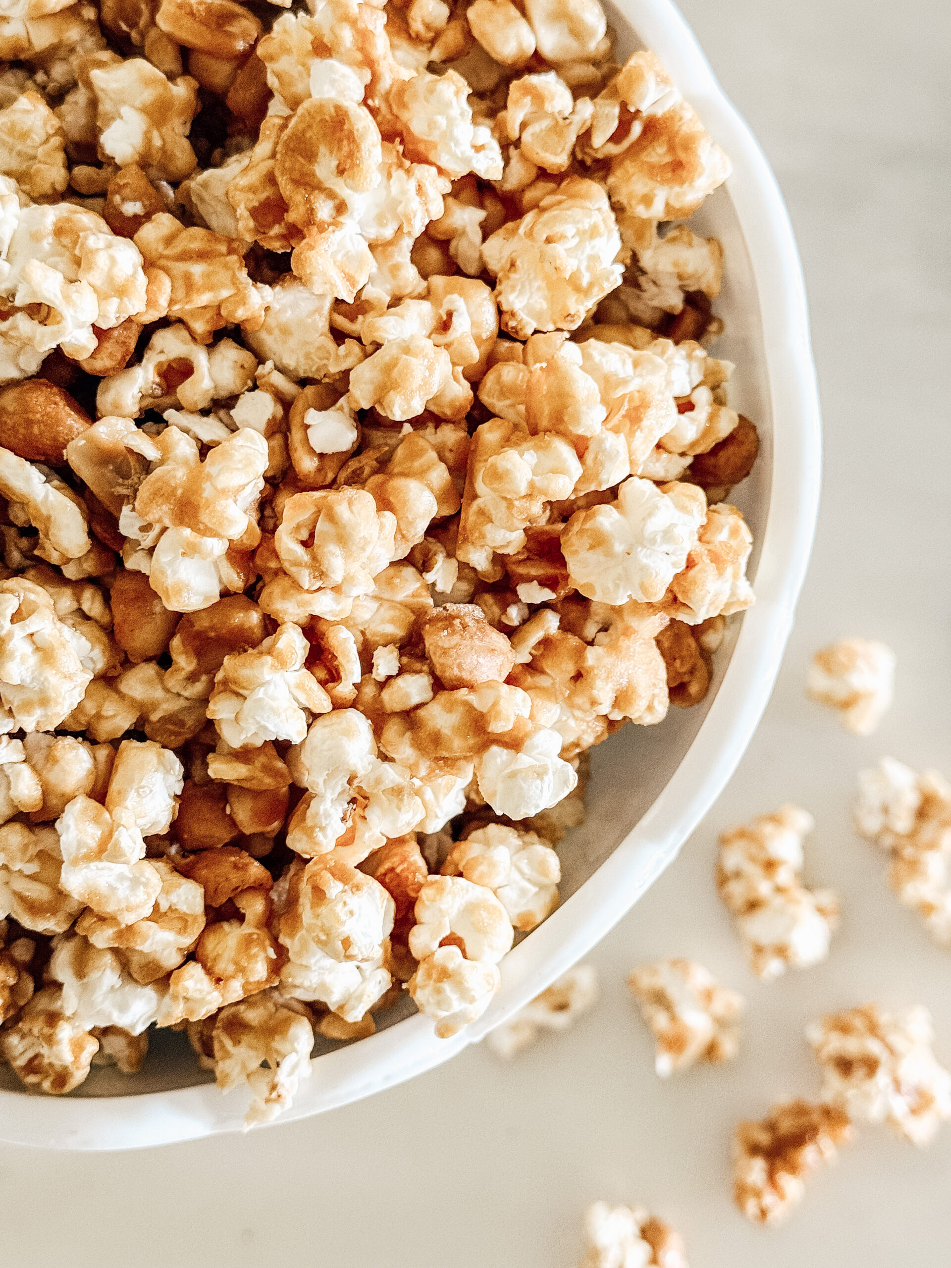 A close-up of a white bowl filled with caramel-coated popcorn, with a few pieces scattered on a light-colored surface beside the bowl.