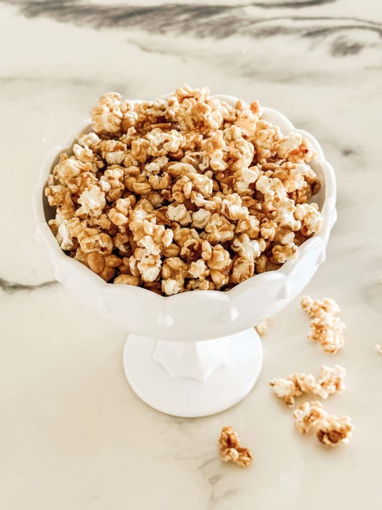 A white pedestal bowl filled with caramel popcorn sits on a marble surface, with a few popcorn pieces scattered nearby.