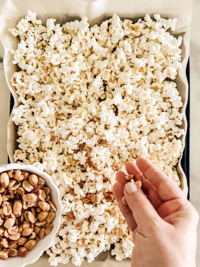 A hand holding a few peanuts over a baking tray filled with popcorn; a small bowl of shelled peanuts sits on the trays edge.