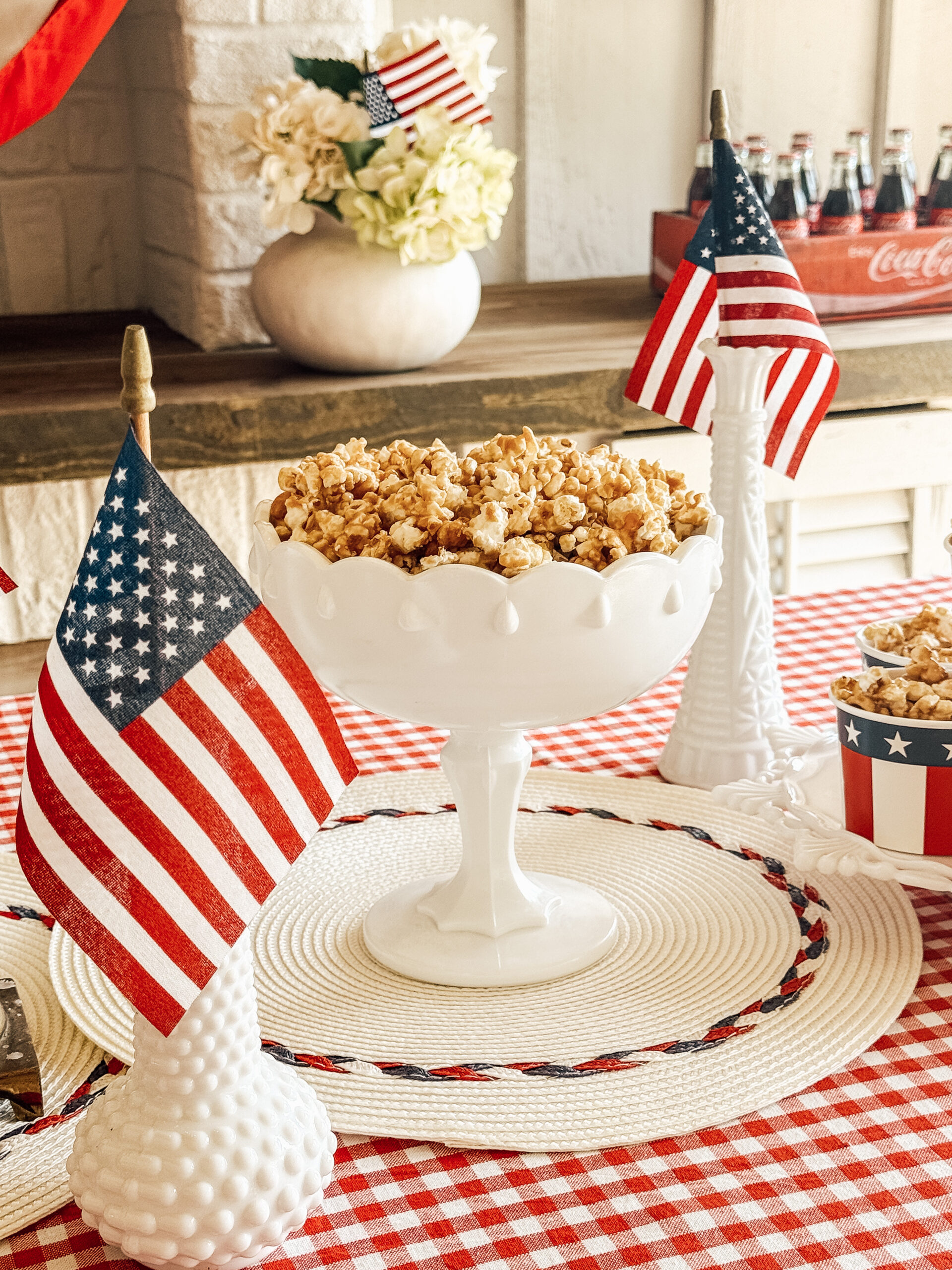 A white pedestal bowl filled with caramel popcorn sits on a red and white checkered tablecloth, decorated with small American flags and white vases, creating a festive patriotic display.