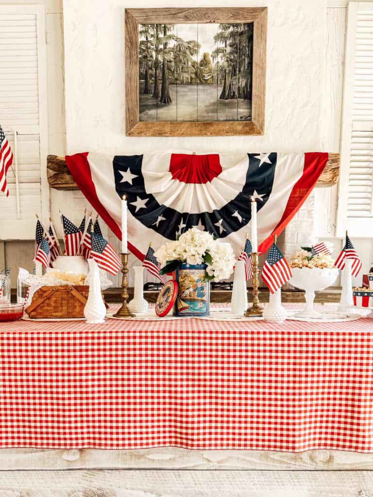 A table with a red and white checkered tablecloth is decorated with American flags, white flowers, candles, and patriotic bunting. A landscape painting hangs on the wall above the display.