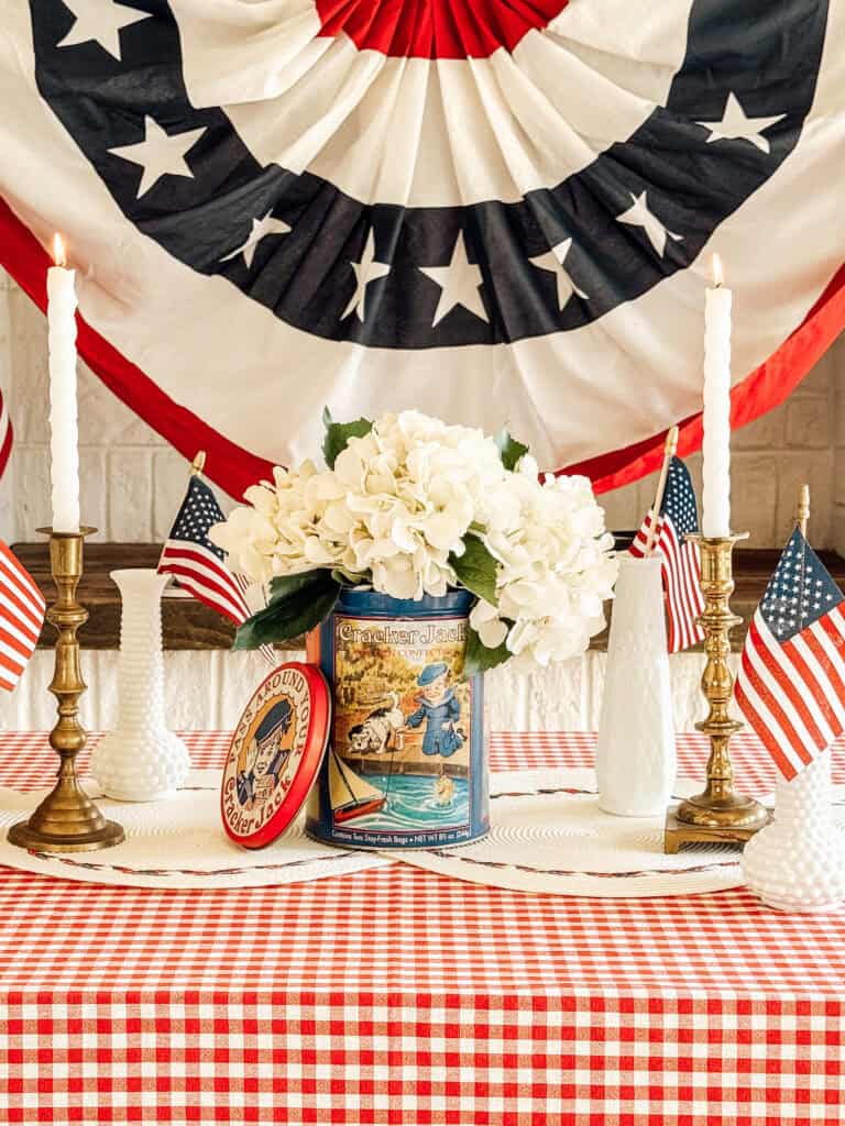 A patriotic table display features white flowers in a vintage tin, brass candlesticks with white candles, small American flags, and a red-and-white checkered tablecloth, set against a backdrop of red, white, and blue bunting.
