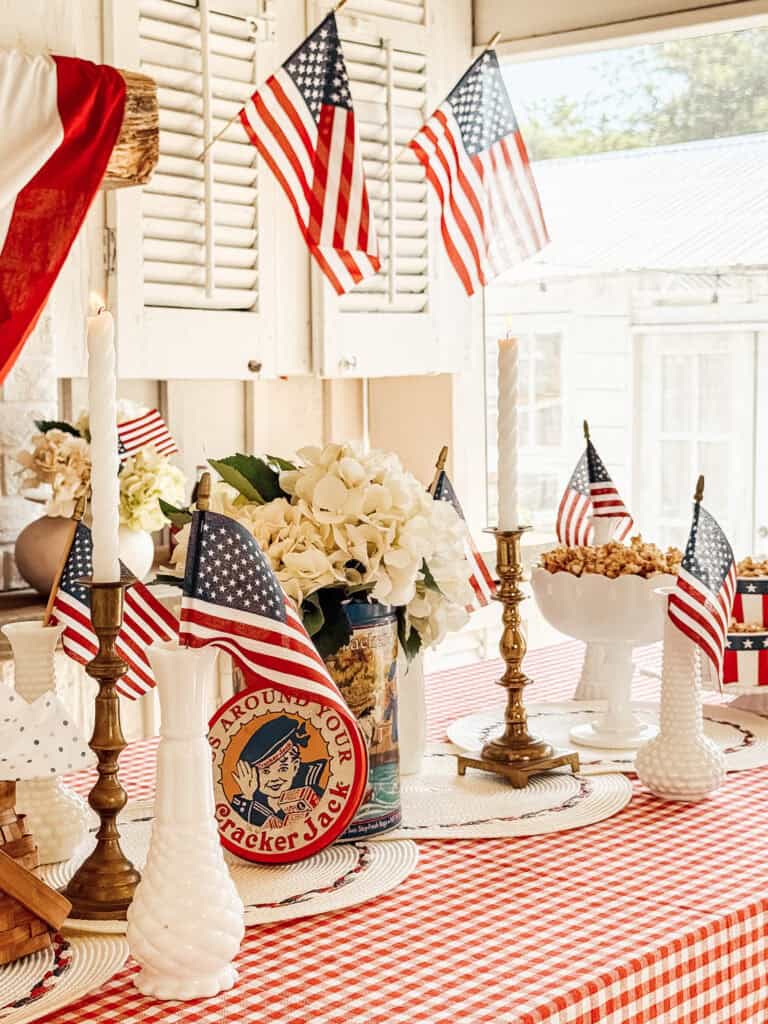 A table with a red and white checkered tablecloth is decorated with American flags, white candlesticks, brass candle holders, white flowers, a vintage Cracker Jack tin, and a bowl of popcorn, set in a sunlit room.