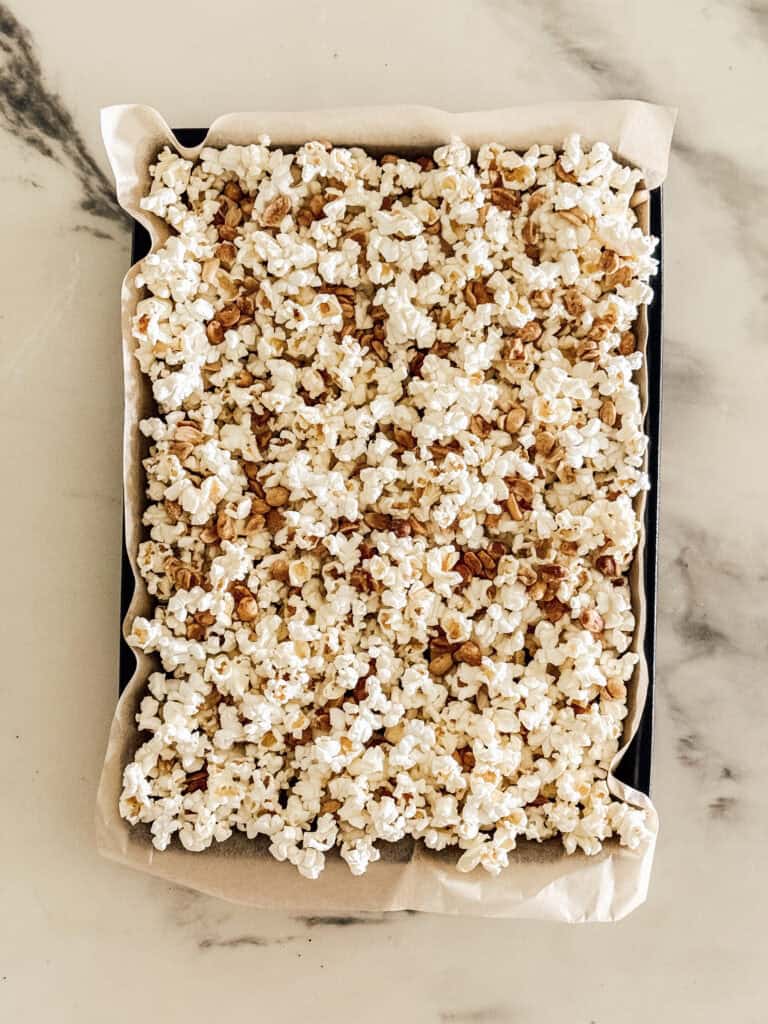A rectangular baking tray lined with parchment paper is filled with homemade caramel popcorn, spread evenly on top of a marble surface.