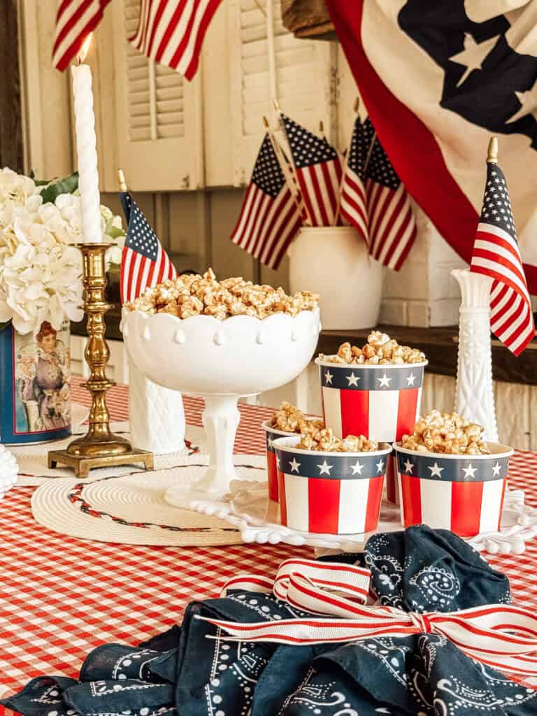 A festive table with caramel popcorn in patriotic red, white, and blue containers, American flags, a white candle in a brass holder, white flowers, and a red checkered tablecloth, decorated for a patriotic celebration.