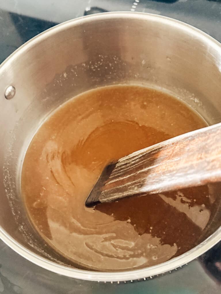 A wooden spoon stirs a golden-brown liquid mixture in a stainless steel pot, likely caramel or sauce, on a stovetop.