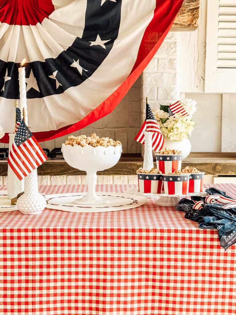 A patriotic table setup with a red and white checkered tablecloth, popcorn in star-spangled cups, American flags, and a large red, white, and blue banner in the background. Candles and bandanas complete the festive decor.
