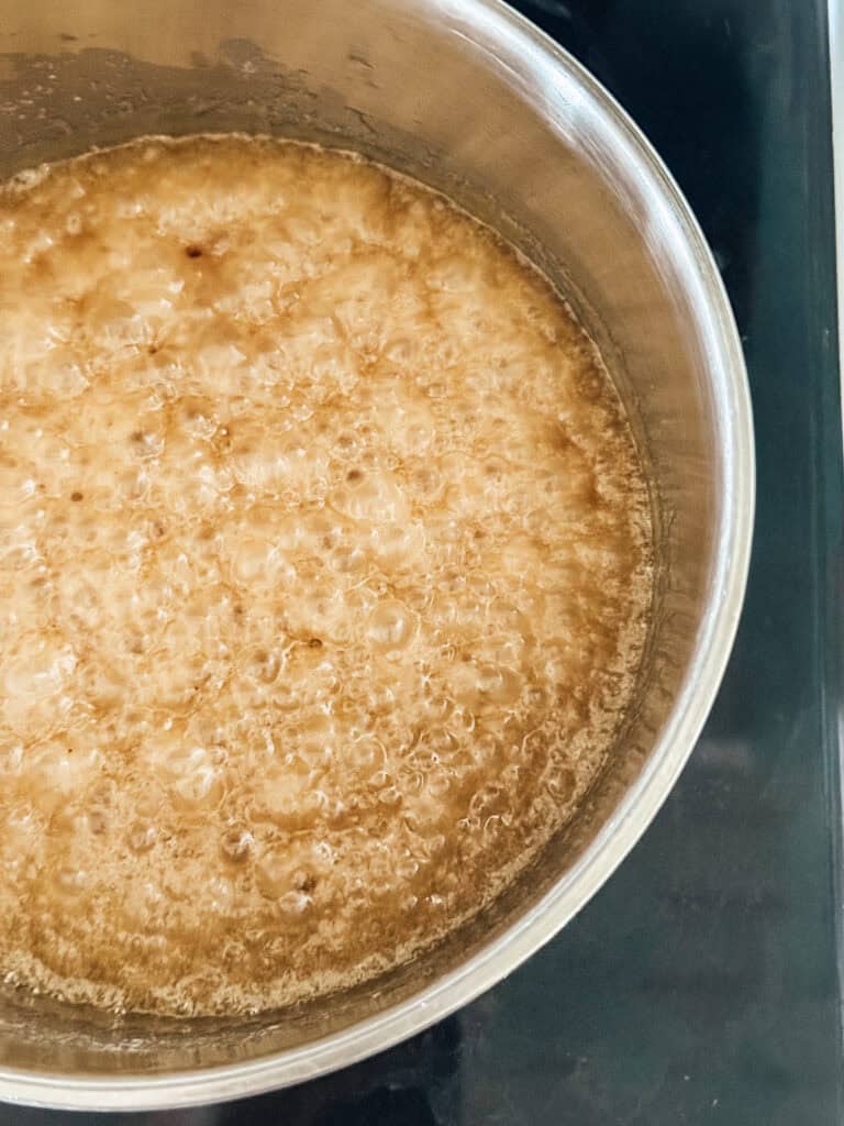 A close-up of caramel sauce bubbling and boiling in a stainless steel pot on a stovetop.