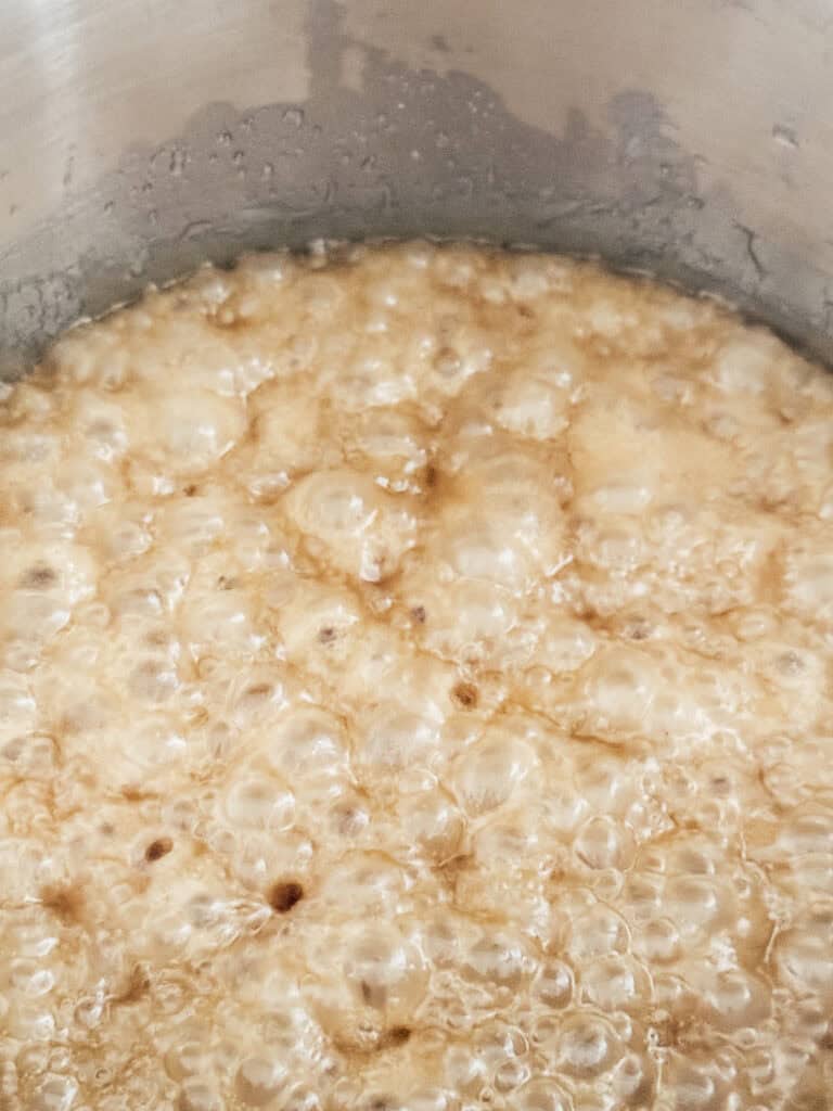 Close-up of bubbling caramel sauce in a stainless steel pot, showing a golden brown color and frothy texture as it cooks.