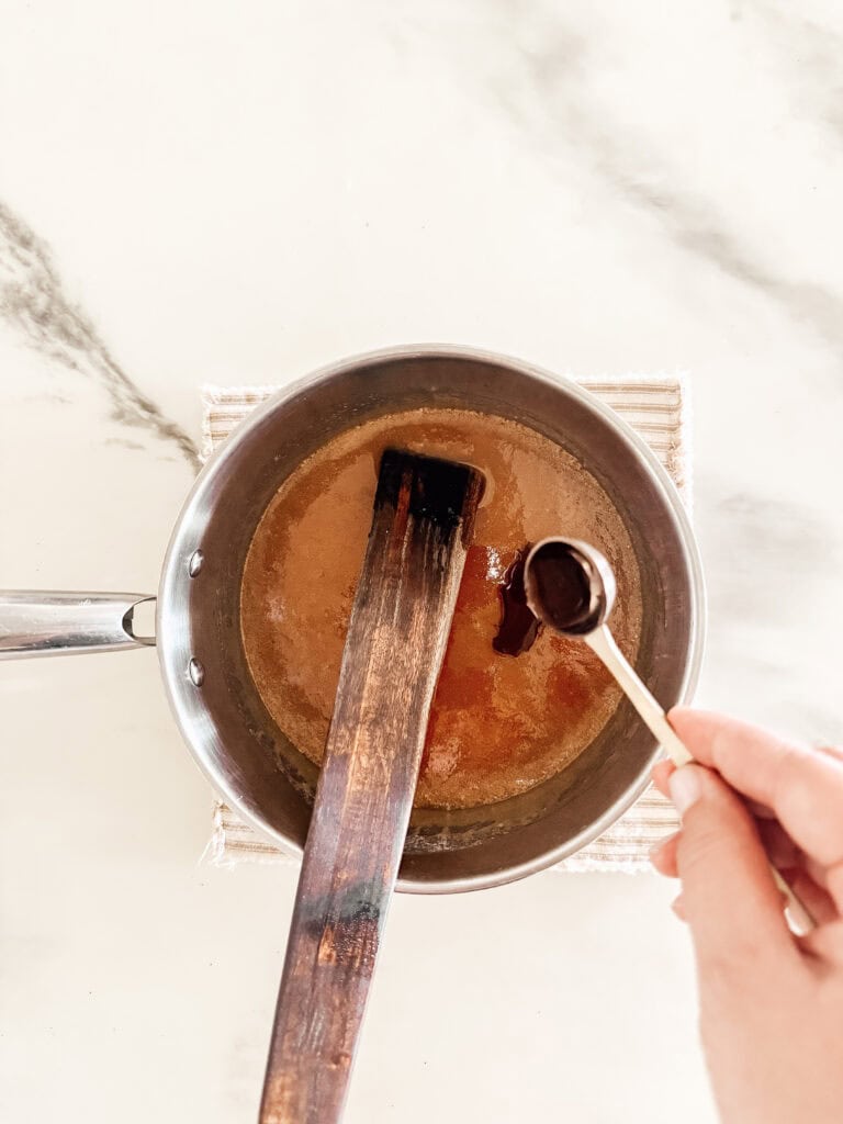 A hand holds a small measuring spoon over a saucepan with a brown liquid, stirring with a wooden spatula on a light countertop.
