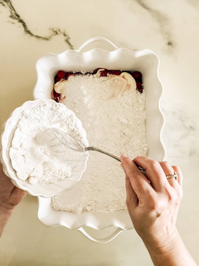 Hands spoon dry cake mix from a bowl into a white baking dish over cherry pie filling and dollops of butter, preparing a dessert on a light countertop.