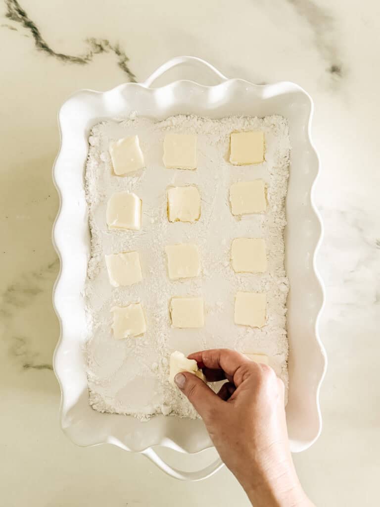 A hand places a slice of butter onto a white baking dish filled with flour. Other butter slices are already evenly spaced on top of the flour.