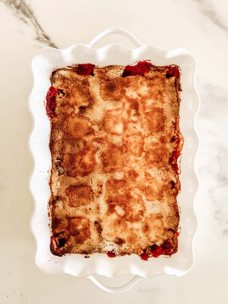A rectangular white baking dish filled with a golden-brown baked cobbler with hints of red fruit showing along the edges, sitting on a light marble surface.