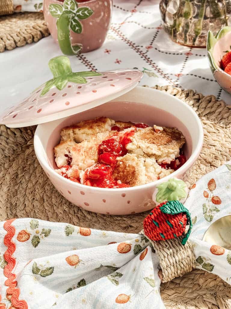 A pink ceramic dish with a green handle holds a golden, baked strawberry dessert. The dish sits on a woven mat, next to a strawberry-patterned napkin and decorative tableware at a picnic setting.